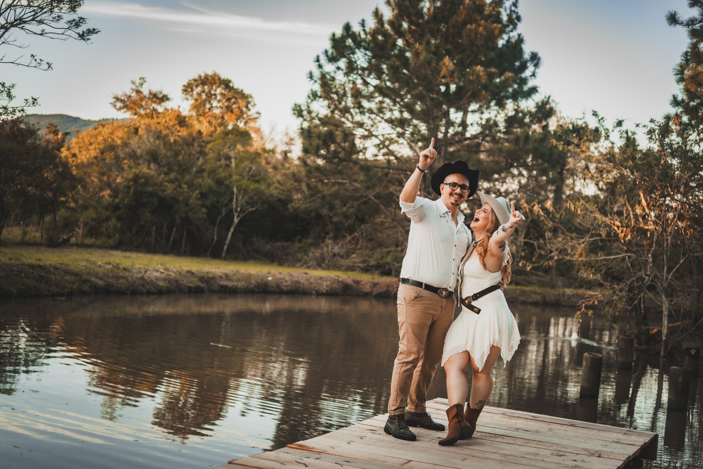 Casal em ensaio ao entardecer, sorrindo e de braços erguidos em um deque à beira do lago