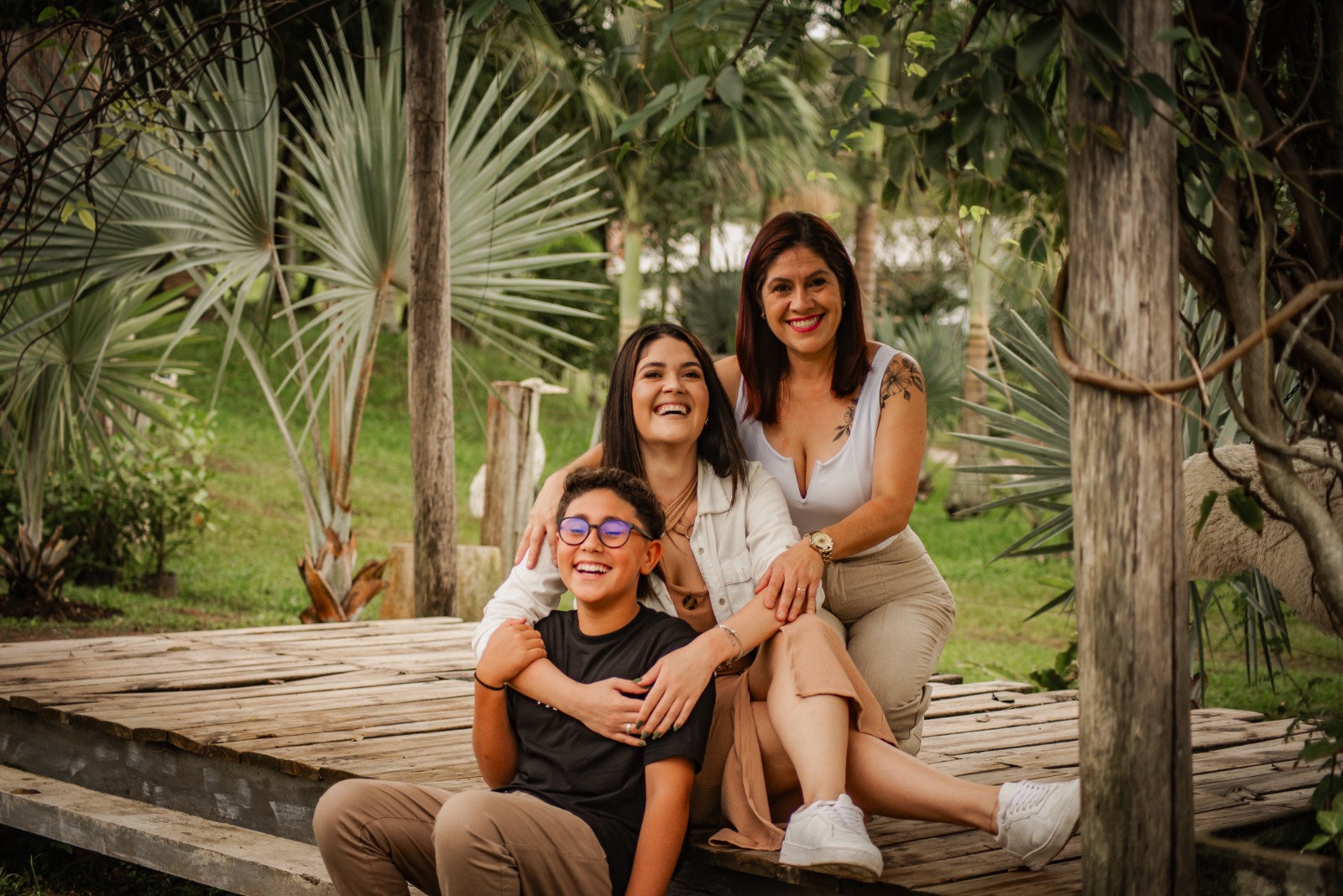 Mãe, filha e filho sorrindo abraçados em um deque entre palmeiras