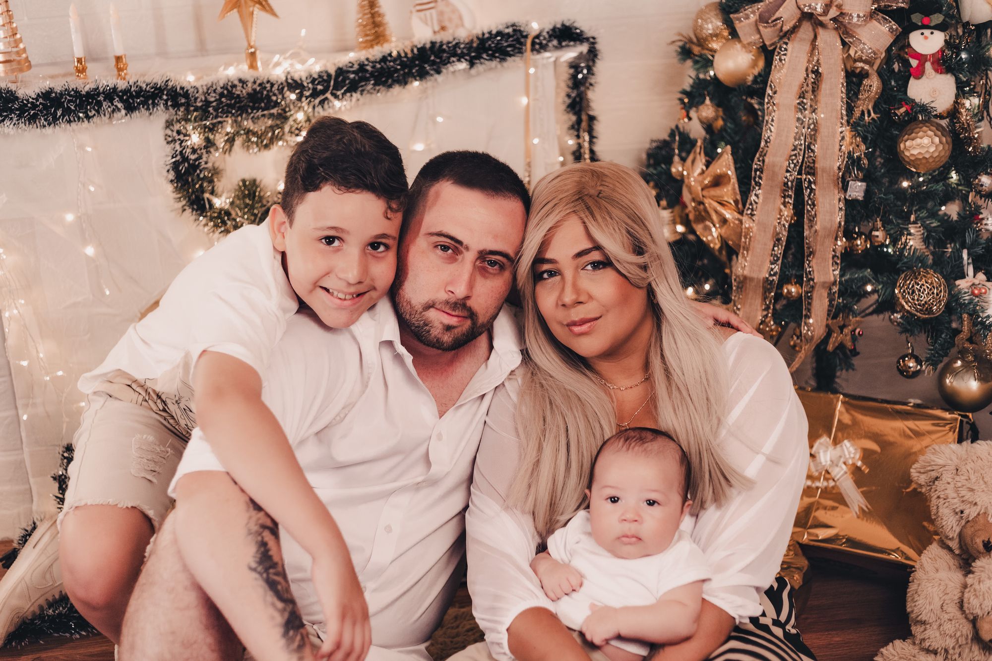 Família com dois filhos, em tons de branco e dourado, diante de uma árvore de Natal com luzes