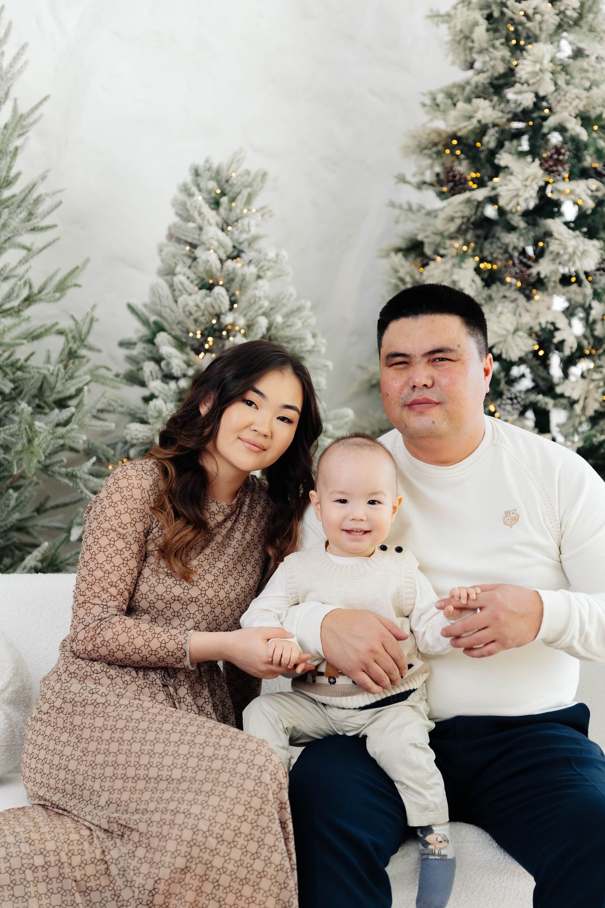 Família sorrindo com o bebê no colo, entre árvores de Natal brancas com luzes douradas