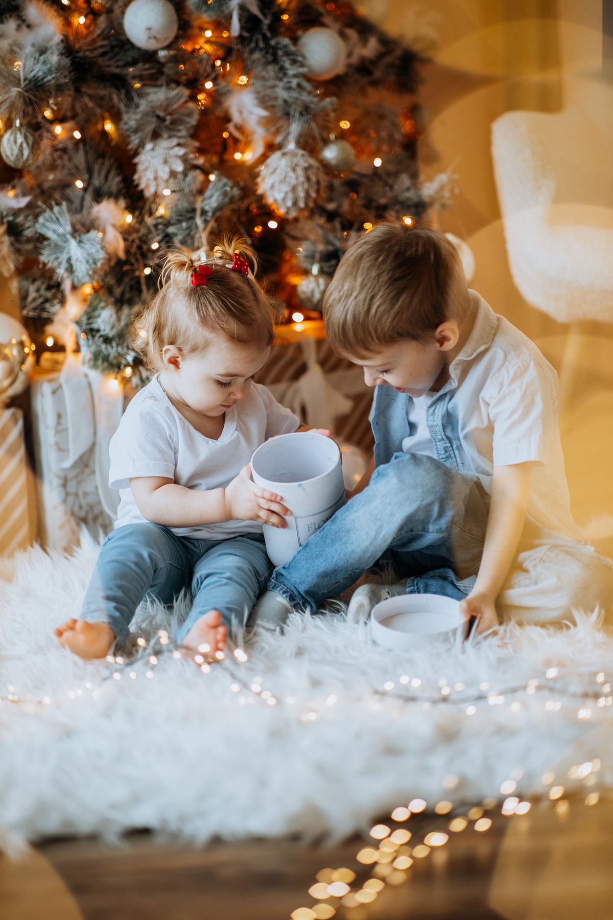 Duas crianças abrindo uma caixa de presente sobre um tapete macio, diante de uma árvore de Natal com luzes quentes